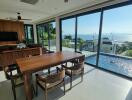 Spacious living area with large glass windows, wooden dining table, chairs, TV on the wall, and a view of the pool and sea.