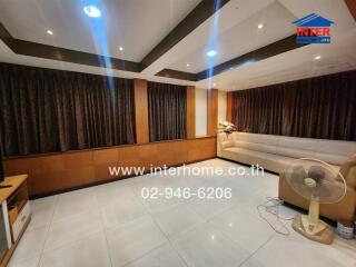 Modern living room with tiled floors, recessed lighting, dark curtains, corner sofa, and a standing fan