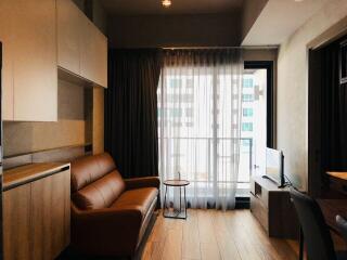 Modern living room with a brown leather sofa, wooden flooring, a small side table, a TV on a cabinet, and large windows with sheer curtains.