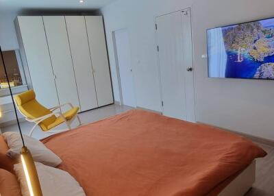 Modern bedroom with a large bed, orange bedding, a TV mounted on the wall, a white wardrobe, and a yellow chair
