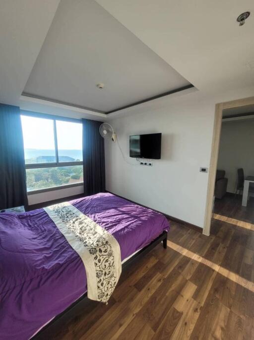 Bedroom with bed, wall-mounted TV, wooden flooring, and large window with curtains