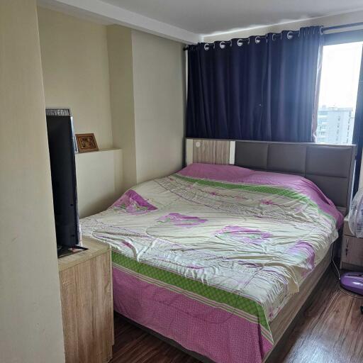 A cozy bedroom with a window and dark curtains, featuring a large bed with colorful bedding and a wooden nightstand holding a television.