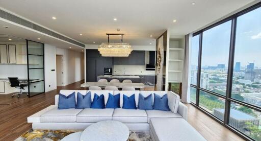 A modern living room with a large gray sectional sofa, decorative pillows, floor-to-ceiling windows, a chandelier, and an open-plan kitchen with a dining area.