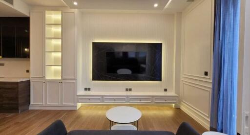 Modern living room with large television, built-in shelves, and hardwood floors