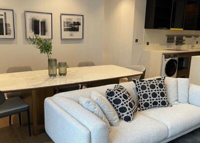 Modern living room with a dining area, including a grey sofa, white marble dining table, wall art, and an open kitchen in the background