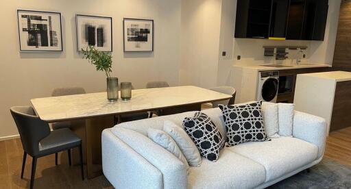 Modern living room with a dining area, including a grey sofa, white marble dining table, wall art, and an open kitchen in the background