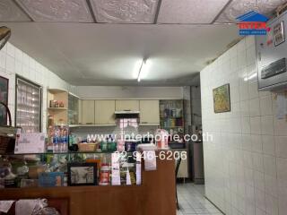 View of a cluttered kitchen space with white tiled walls, wooden cabinetry, and various household items.