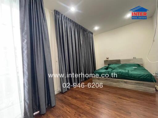 Bedroom with wooden flooring, green bedspread, and large window with gray curtains