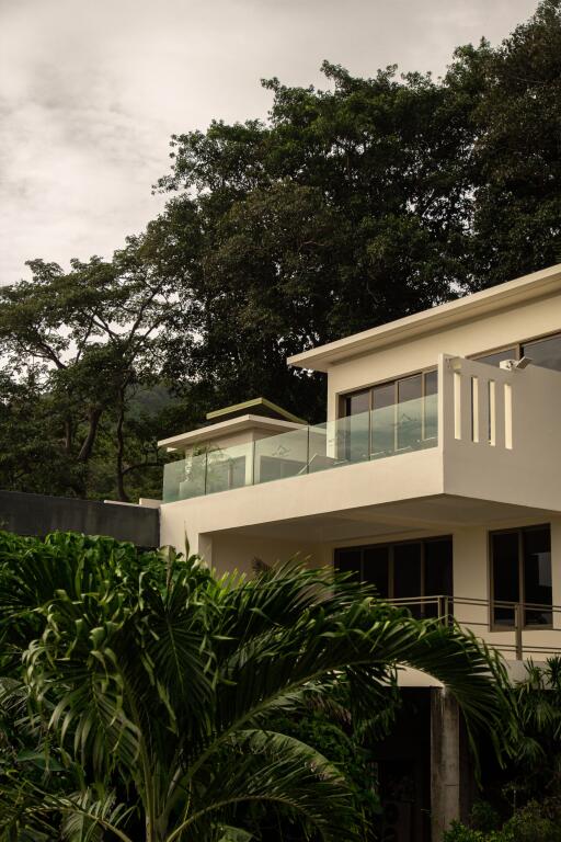 Modern multi-story building with large windows and glass balconies surrounded by lush greenery