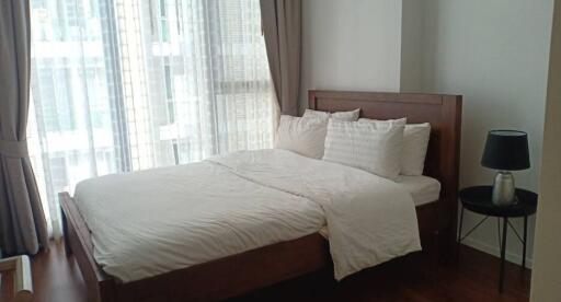 Bedroom with large bed and wooden frame, bedside table with lamp, and large window with curtains