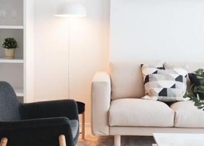 Modern living room with light-colored sofa and decorative pillows, black accent chair, bookshelf, and potted plant.