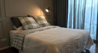 A cozy bedroom with a bed, pillows, lamp, and floor-to-ceiling windows with curtains.