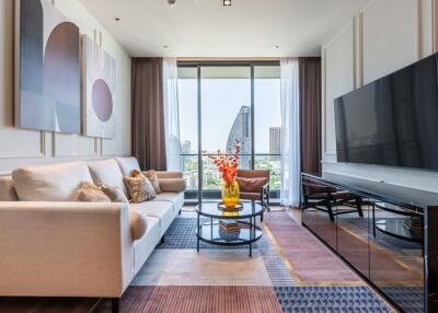 Modern living room with a sofa, large TV, coffee table, decorative items, and a view of tall buildings through large windows