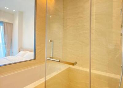 Modern bathroom with glass shower, beige tiles, and a view into a well-lit bedroom