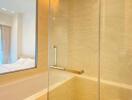 Modern bathroom with glass shower, beige tiles, and a view into a well-lit bedroom