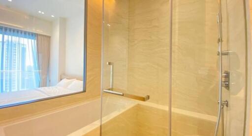 Modern bathroom with glass shower, beige tiles, and a view into a well-lit bedroom