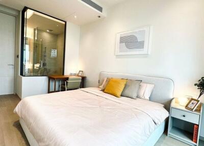 Well-lit bedroom with minimalist decor, featuring a bed, bedside table, desk, and a glass-enclosed bathroom space.