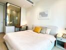 Well-lit bedroom with minimalist decor, featuring a bed, bedside table, desk, and a glass-enclosed bathroom space.