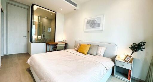 Well-lit bedroom with minimalist decor, featuring a bed, bedside table, desk, and a glass-enclosed bathroom space.