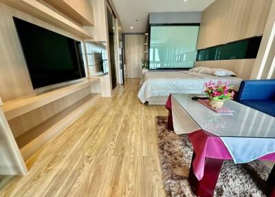 Modern bedroom with a bed, wall-mounted TV, glass partition, wood flooring, and a coffee table with decorative items.