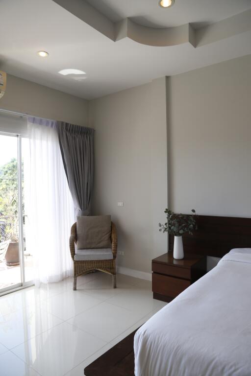 A modern bedroom with a large window, white curtains, and gray walls featuring a bed, bedside table with a vase, and a wicker chair.