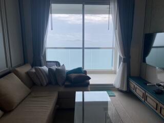 Living room with a sectional sofa, glass coffee table, wall-mounted TV, and a view of the sea through a large window.