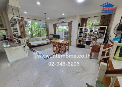 Spacious and well-lit living room with a view to the garden through large windows, featuring a cozy sitting area, dining table, bookshelf, and modern kitchen.