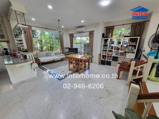 Spacious and well-lit living room with a view to the garden through large windows, featuring a cozy sitting area, dining table, bookshelf, and modern kitchen.