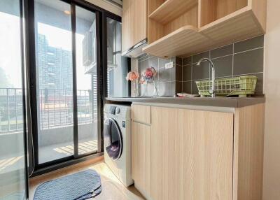Modern kitchen area with light wood cabinets, a washing machine, and a small balcony.
