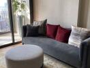 Modern living room with a large window, gray sofa, round ottoman, and decorative cushions