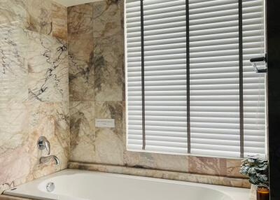 Modern bathroom with marble tiles and a bathtub next to a window with blinds.