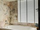 Modern bathroom with marble tiles and a bathtub next to a window with blinds.