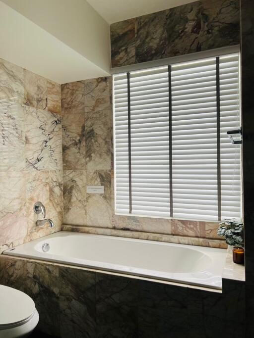 Modern bathroom with marble tiles and a bathtub next to a window with blinds.