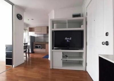 A cozy living area with a TV, shelves, and a view into the kitchen.