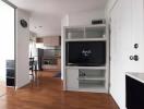 A cozy living area with a TV, shelves, and a view into the kitchen.