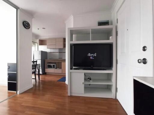 A cozy living area with a TV, shelves, and a view into the kitchen.