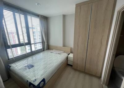 A modern bedroom with a large window, wooden wardrobe, and a mattress on a wooden bed frame.