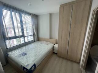 A modern bedroom with a large window, wooden wardrobe, and a mattress on a wooden bed frame.