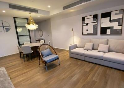 Modern living room with wooden floor, large sofa, decorative art on the wall, a dining table with chairs, and a contemporary chandelier.