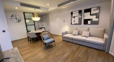 Modern living room with wooden floor, large sofa, decorative art on the wall, a dining table with chairs, and a contemporary chandelier.