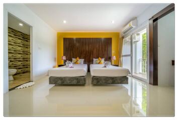 Modern bedroom with two beds and a yellow accent wall, adjacent bathroom, and a sliding door leading to a balcony.