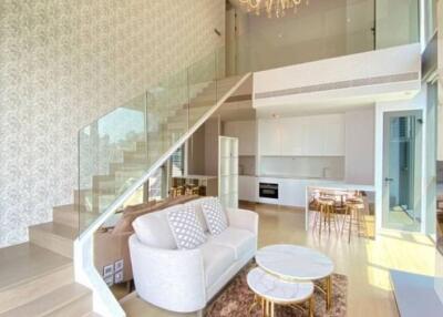 Modern duplex living area with white sofa, glass railing staircase, chandelier, and open kitchen