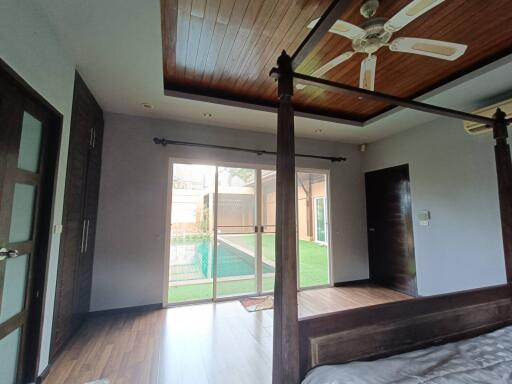 Spacious bedroom with wooden ceiling and four-poster bed, overlooking a garden with a swimming pool.