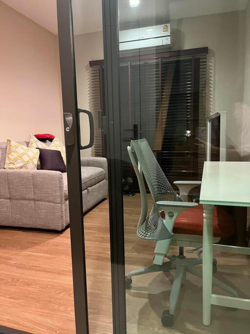 Glass partition separating a study area with a desk and ergonomic chair from a living room with a sofa and decorative pillows.