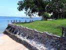 Beachfront property with a stone retaining wall, grassy lawn, and lush trees