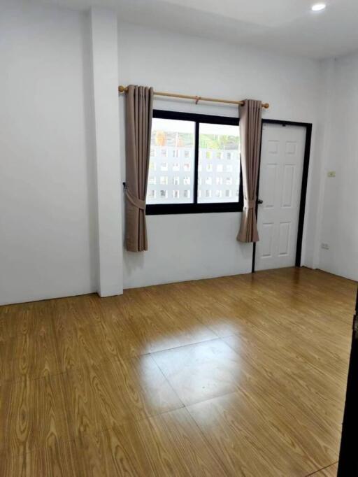 Empty bedroom with wooden flooring, a window with curtains, and a door.