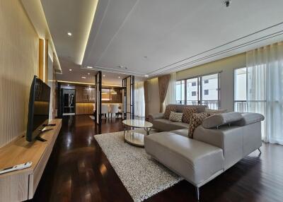 Modern living room with ample natural light, sectional sofa, TV, and open-plan kitchen.