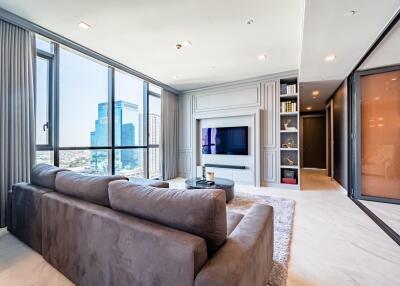 Modern living room with floor-to-ceiling windows, large gray couch, TV, built-in shelves, and city view.