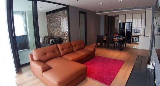 Modern living room with an open kitchen, featuring a brown sectional sofa, red rug, open dining area, and stylish decor