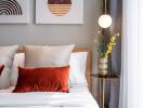 Cozy bedroom with modern decor, featuring a bed with white and orange pillows, a bedside table with a lamp, and artwork on the wall.